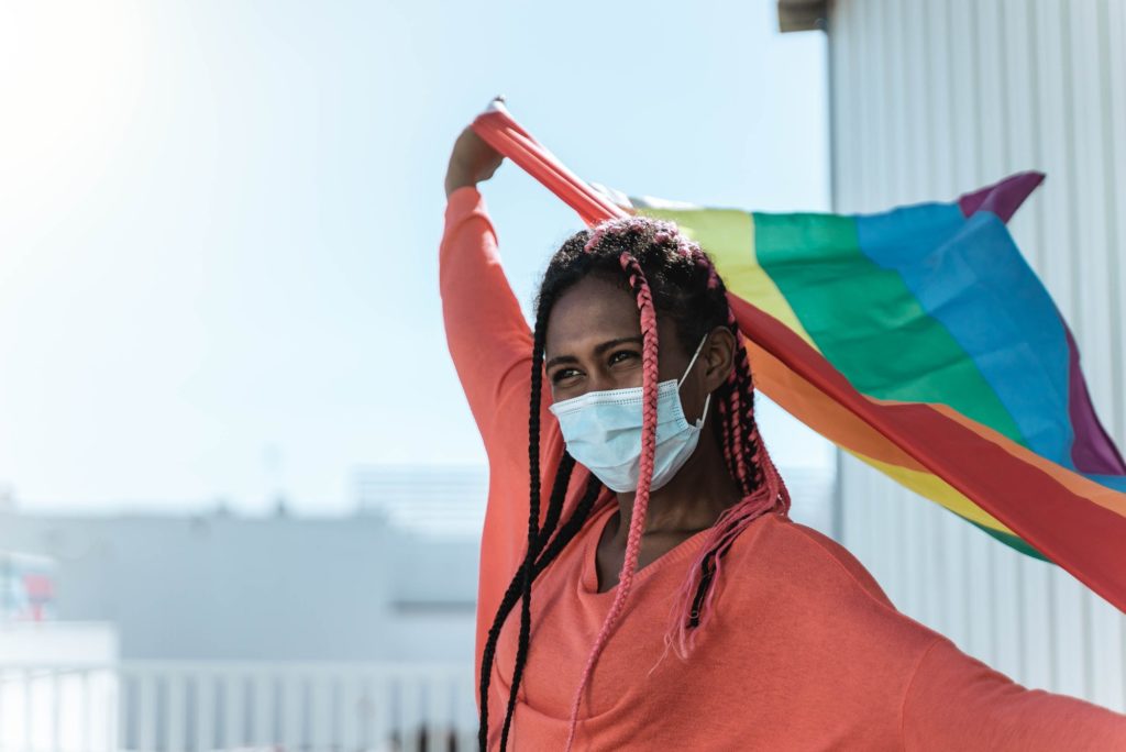 Young african lesbian with safety mask holding lgbt rainbow flag at pride event - Focus on face