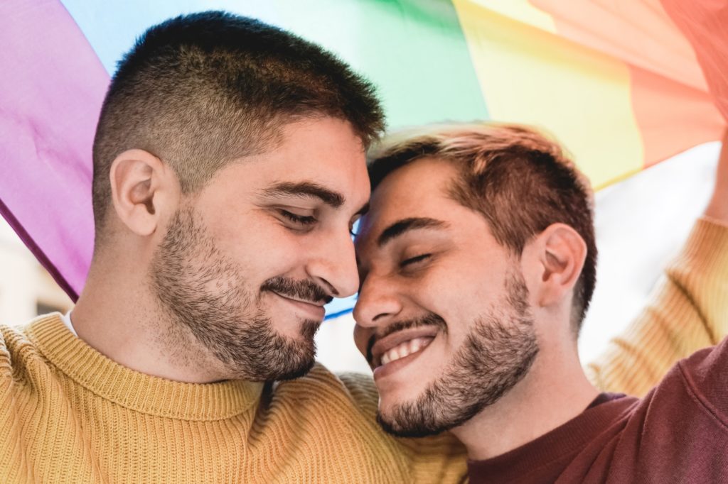 Young gay men couple having tender moment under rainbow LGBT flag outdoor - LGBTQ+ love concept